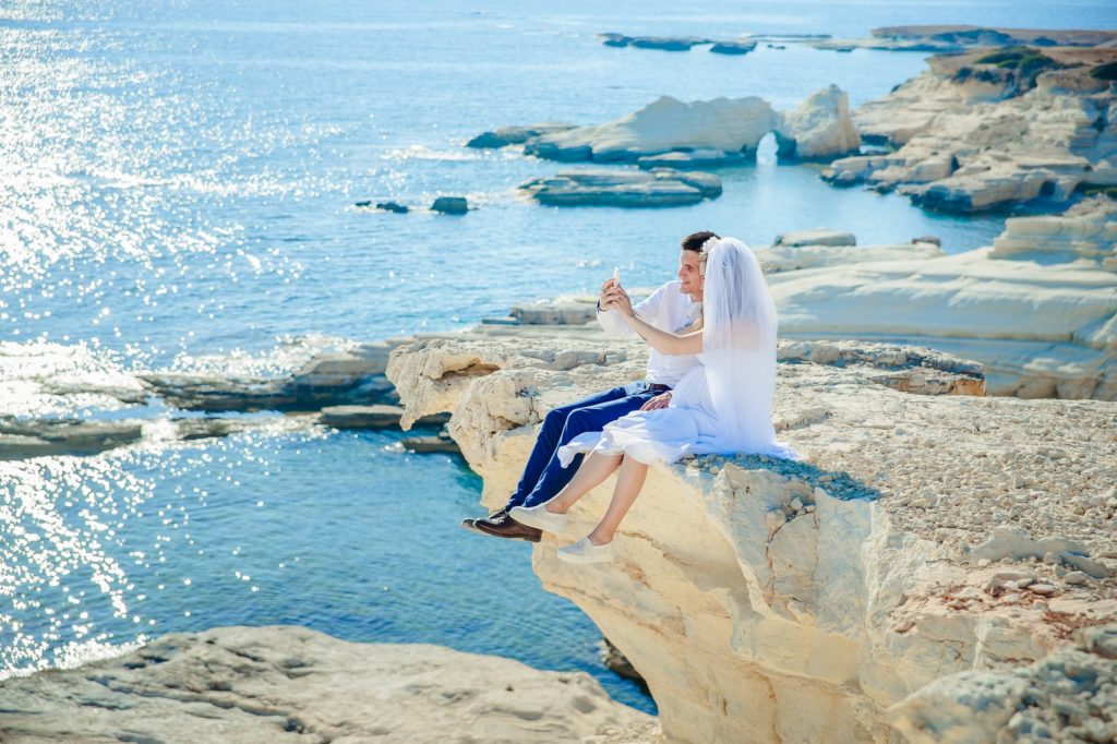woman wearing wedding dress sitting beside man wearing white dress shirt on the stone tip facing the sea